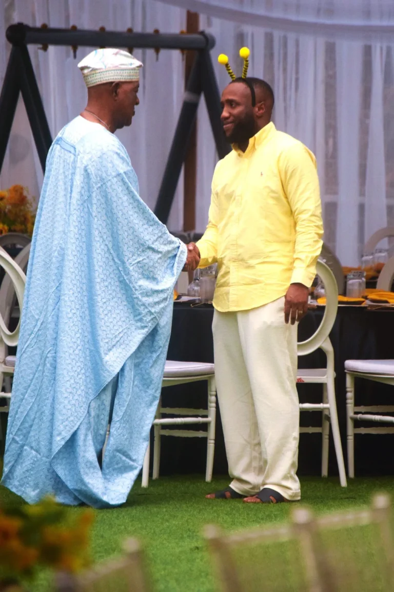 Man in yellow shirt with bee antennae headband shakes hands with elder in blue agbada.