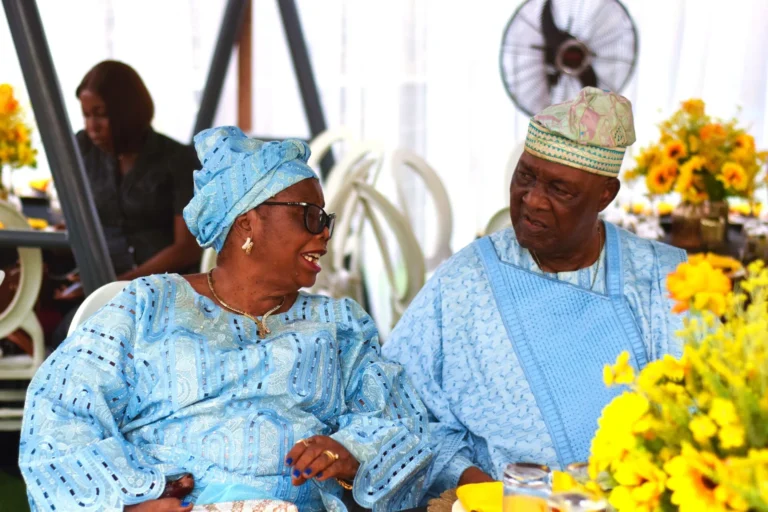 Older couple in matching pale-blue Nigerian attire share a fond conversation at a table.