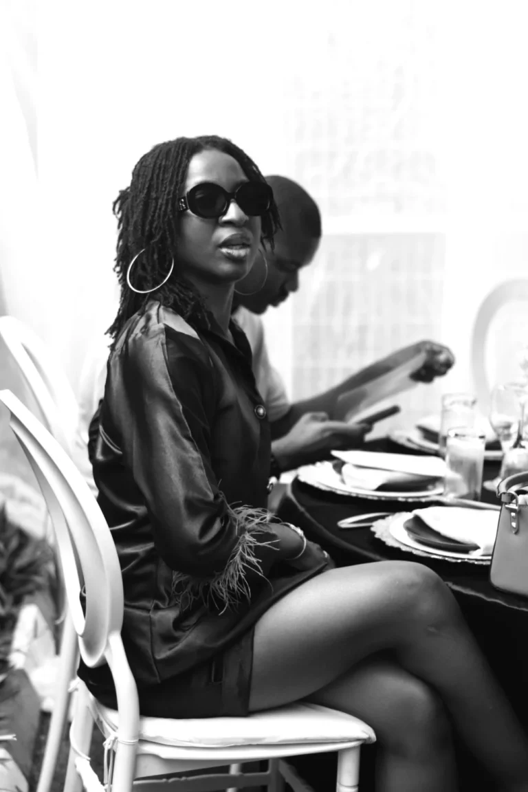 Monochrome portrait of woman in feathered dress and hoop earrings at a dining table.