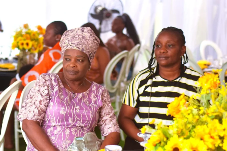Two women in patterned dresses sit beside sunflowers at a marquee gathering.