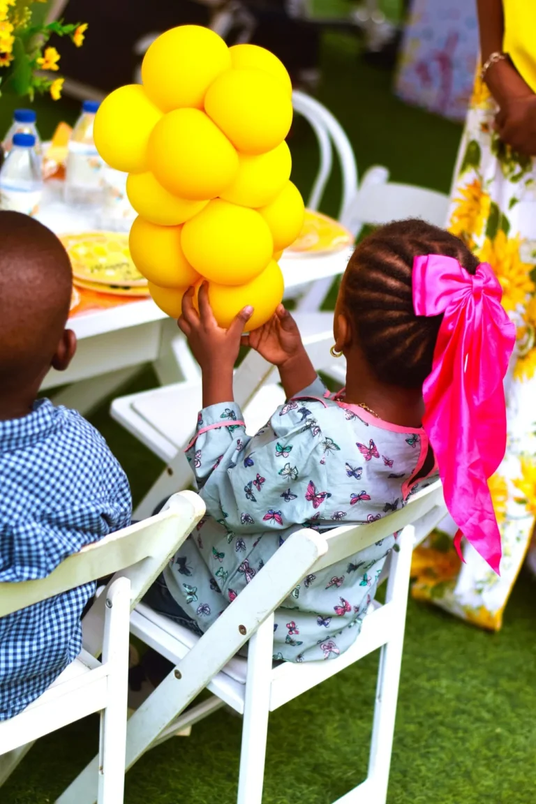 Children seen from behind hold a yellow balloon cluster while seated on white chairs.