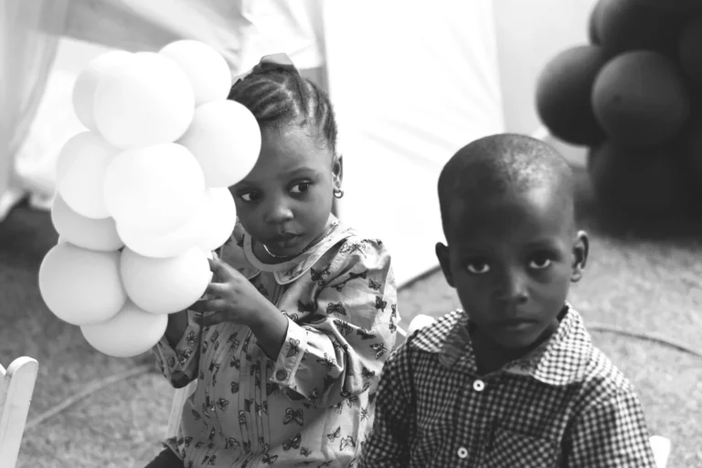 Monochrome image of two small children, one holding a balloon cluster at a party.