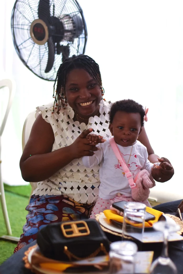 Smiling woman with braids holds a baby in a pink bag in front of an electric fan.