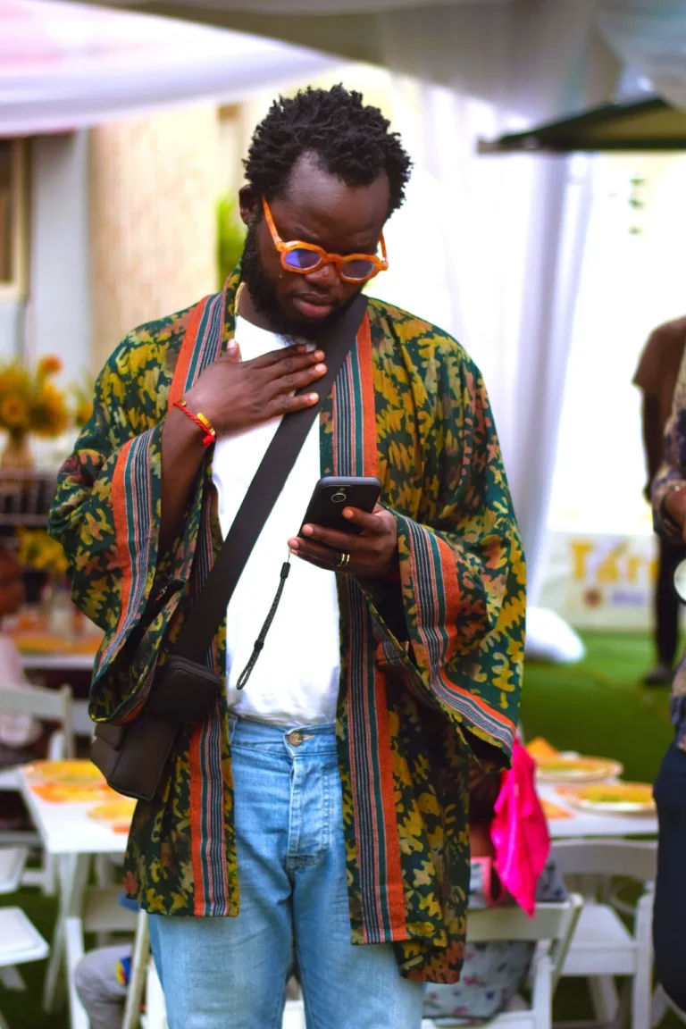 Man in patterned kimono and orange sunglasses checks his phone at an outdoor event.