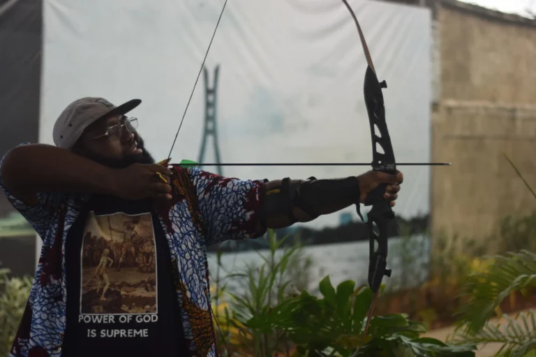 Bearded man in patterned shirt draws back a black recurve bow, aiming towards Lagos's Lekki bridge.