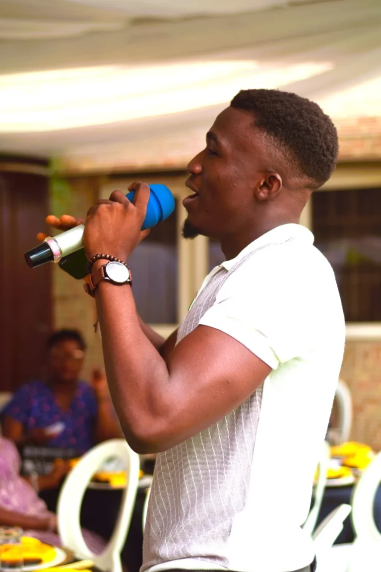 Young man in white polo shirt sings into a blue microphone at an indoor gathering.