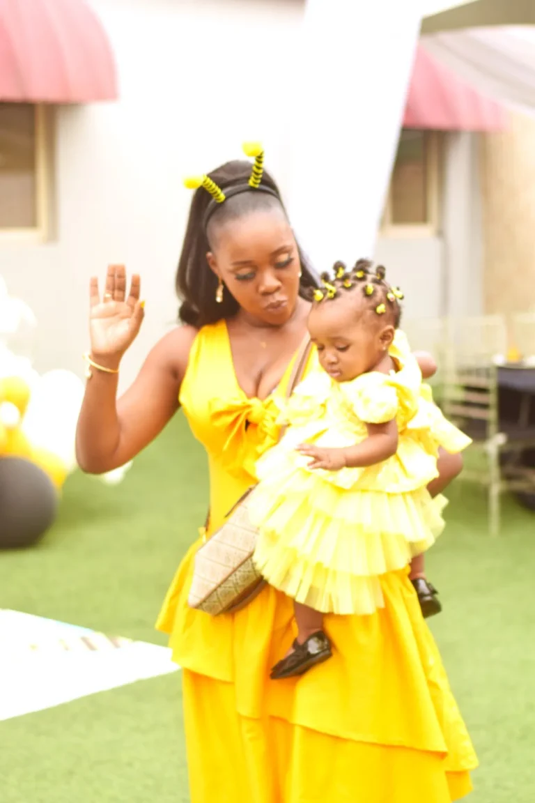 Woman in yellow dress holds a baby girl in matching yellow ruffled dress outdoors.