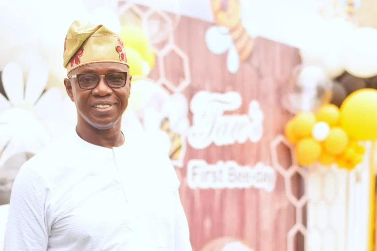 Smiling older man in white tunic and traditional cap stands by a honeycomb-themed backdrop.