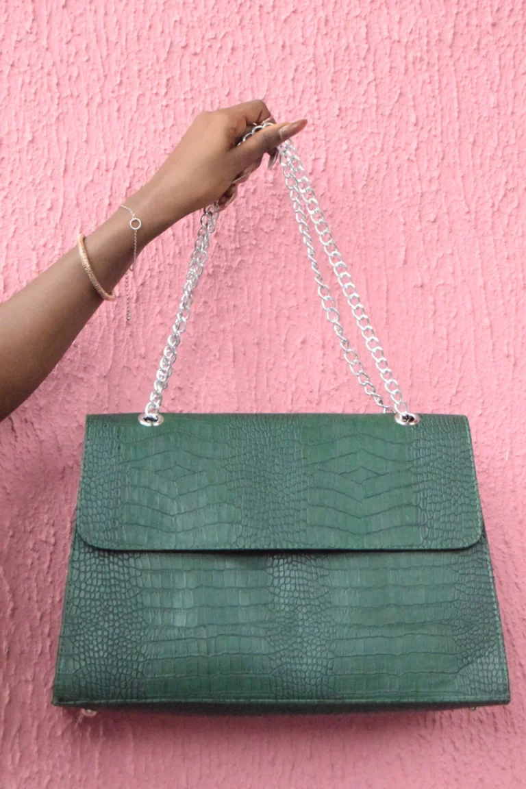 Hand holds up a green crocodile-embossed handbag with silver chain straps against a pink wall.