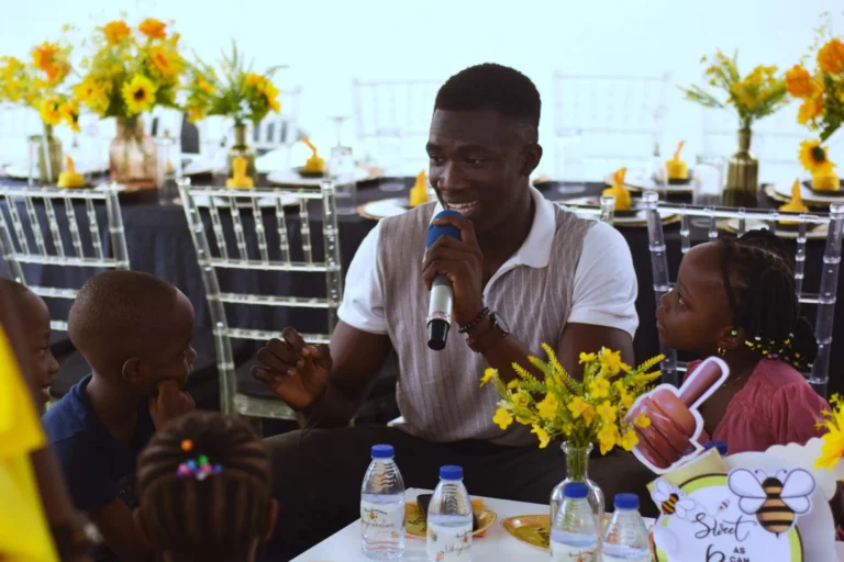 Young man holds a microphone speaking to seated children at a sunflower-decorated table.
