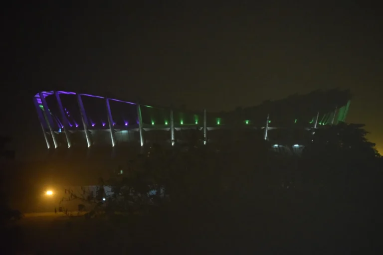 Misty night view of a curved stadium lit with purple and green lights beneath a dark sky.