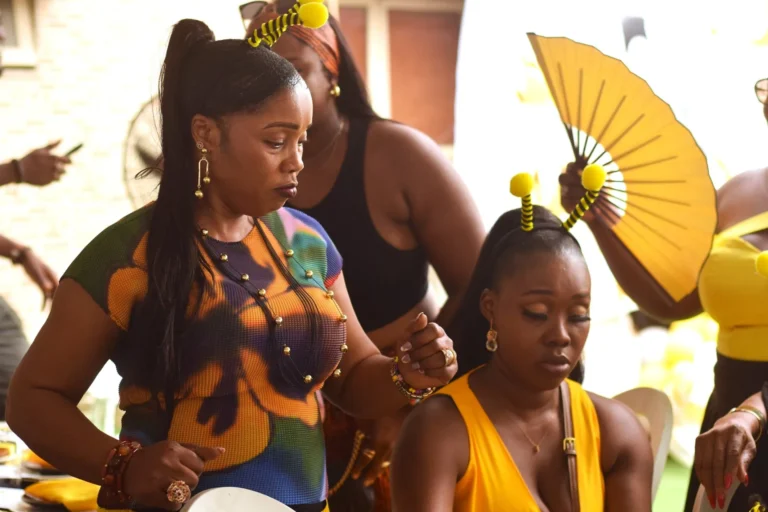Three women in colourful outfits stand together with a yellow hand fan raised behind them.