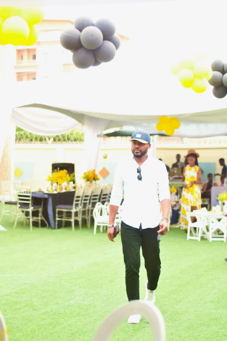 Man in white shirt and cap walks across green turf beneath yellow and grey balloon clusters.