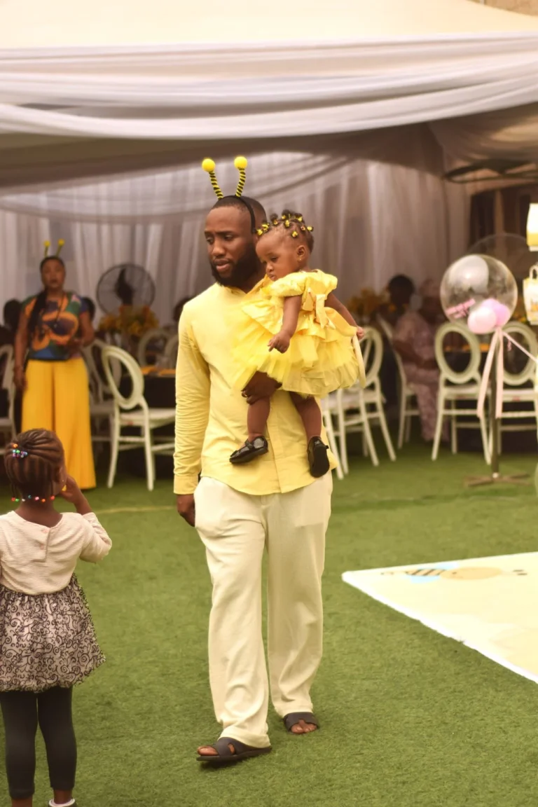 Father with bee antennae headband carries his young daughter in a yellow ruffled dress.