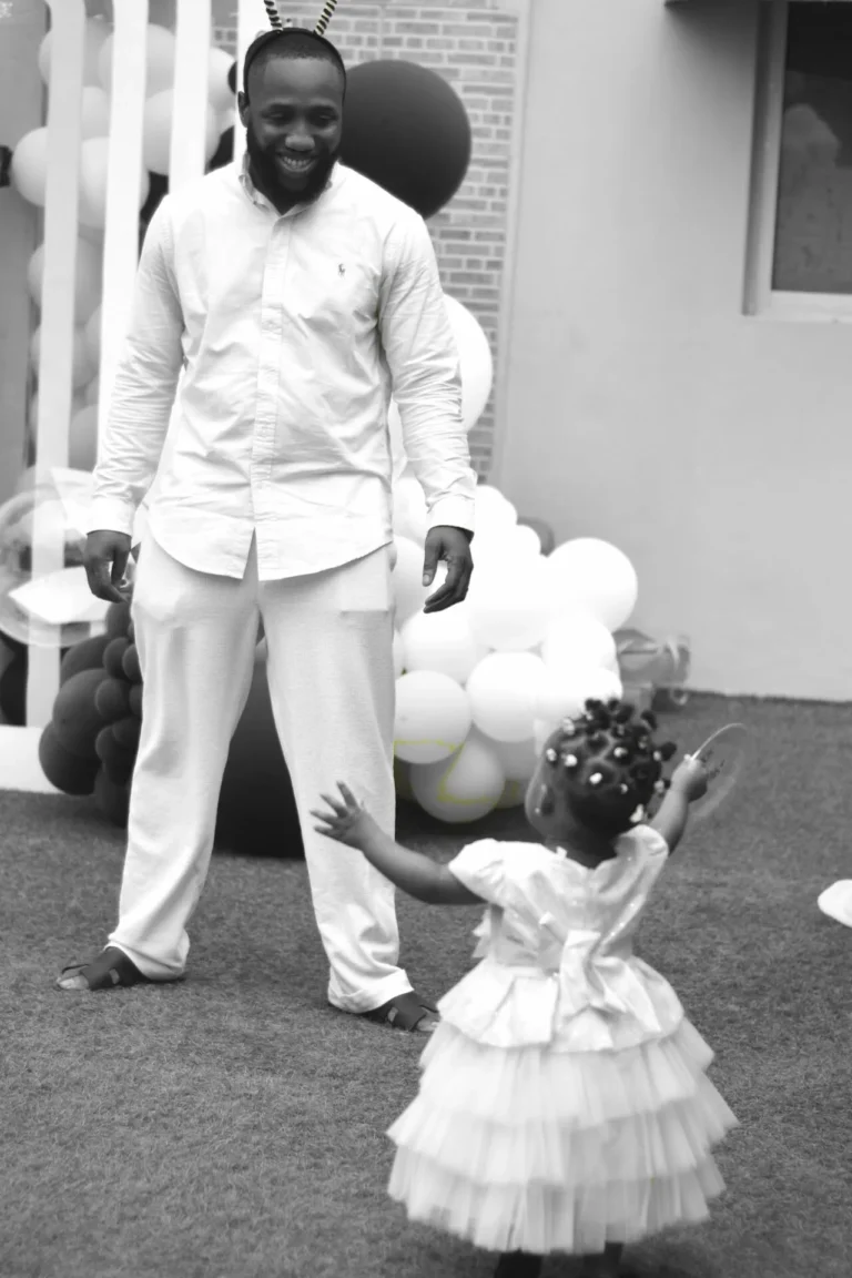Bearded man smiles down at his small daughter in a tutu dress, in black and white.