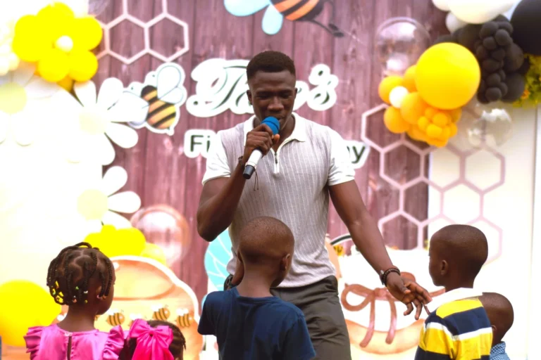 Man sings into a microphone surrounded by young children at a bee-themed party backdrop.