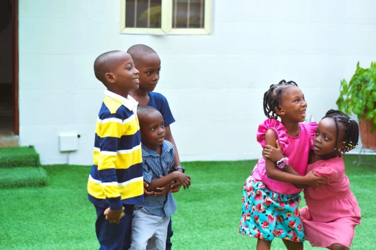 Five children laugh and play together on artificial grass beside a white wall.