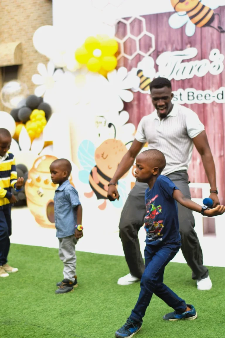 Boys dance with an adult in front of a bee-themed birthday backdrop on green turf.