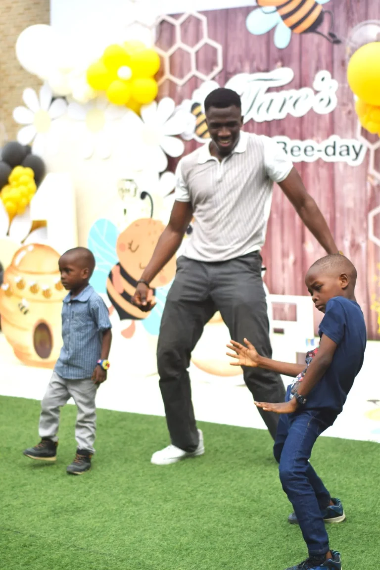 Man and young boy share a dance move on green turf at a child's bee-themed party.