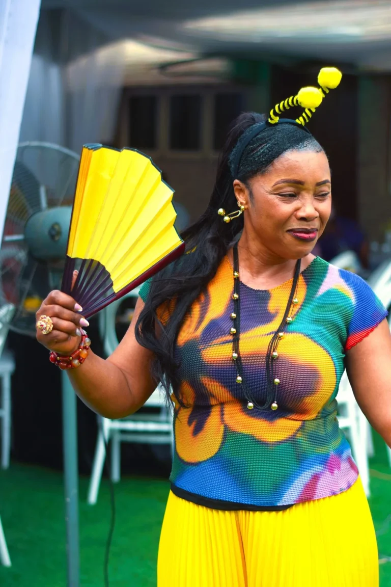 Woman with bee antennae headband holds a yellow fan, wearing a vivid floral top.