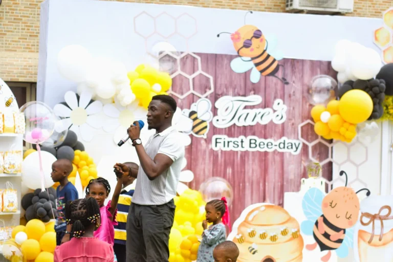 Young man speaks into a microphone among children at a bee-themed birthday backdrop.