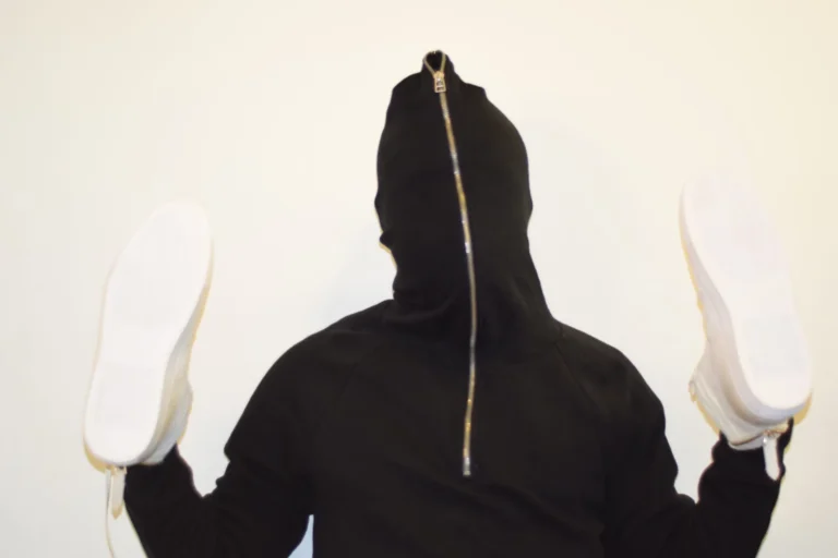 Person in black hooded top holds two white trainers aloft, hood zipped over their face.