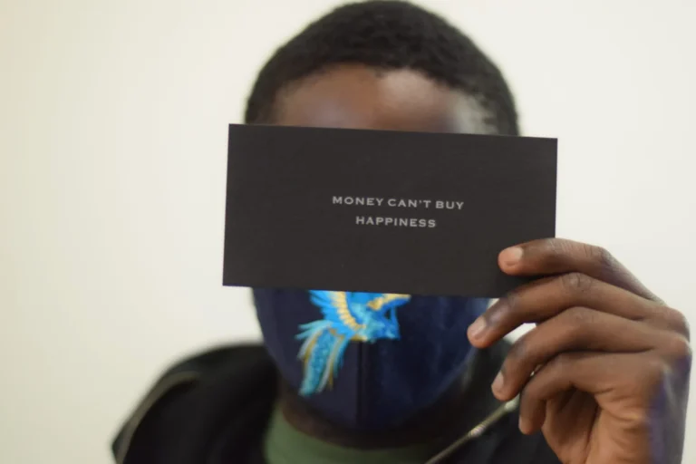 Person in patterned face mask holds a black card reading 'Money can't buy happiness' to the camera.