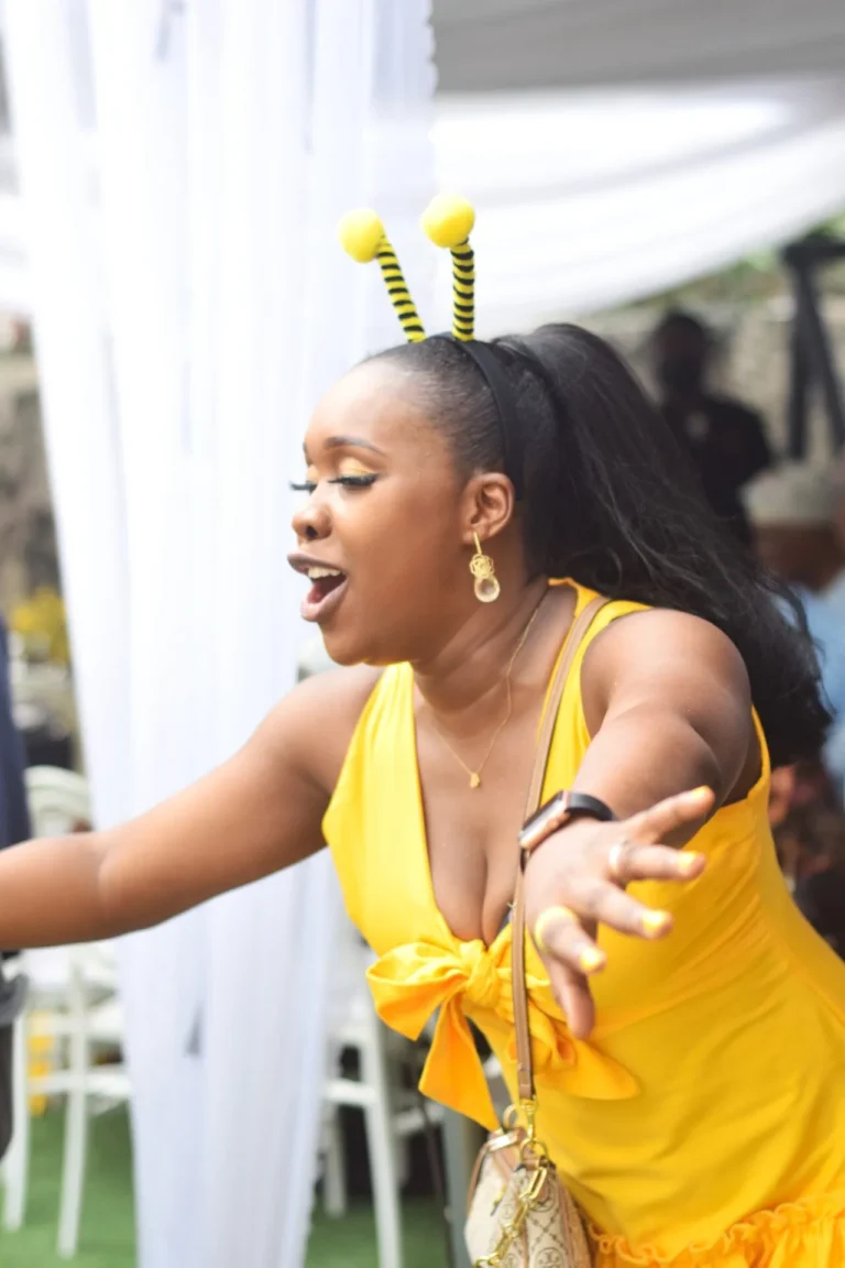 Woman with bee antennae headband in yellow dress reaches out mid-dance, mouth open in delight.