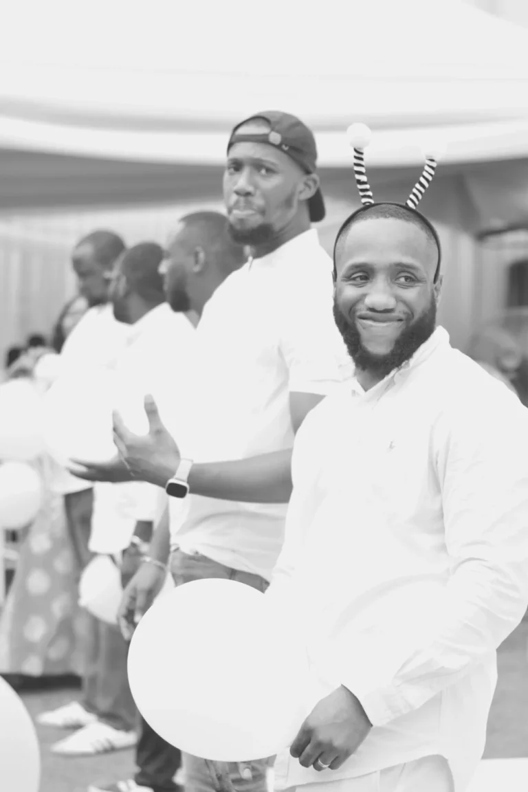 Men in white shirts queue with white round balloons, one wearing striped bee antennae.