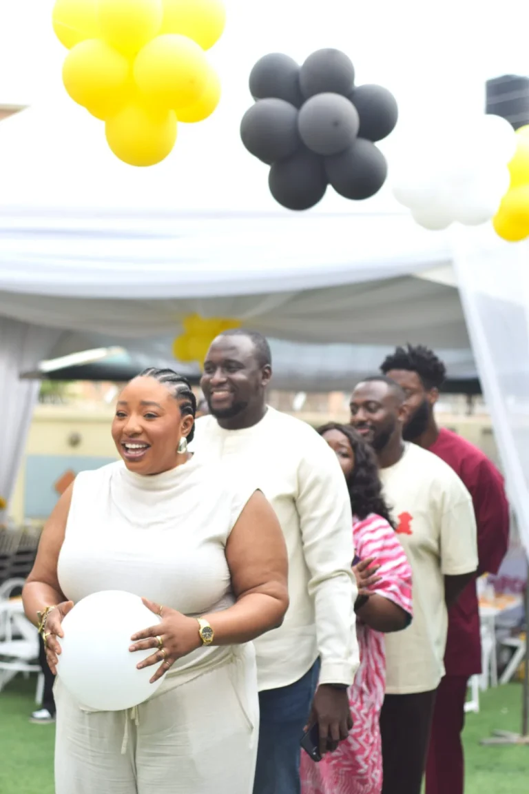 Group of smiling guests line up under a marquee beneath yellow and grey balloon clusters.