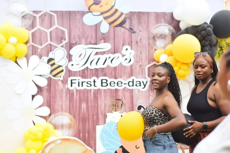 Two young women pose by a wooden Tare's First Bee-day backdrop with balloon decor.