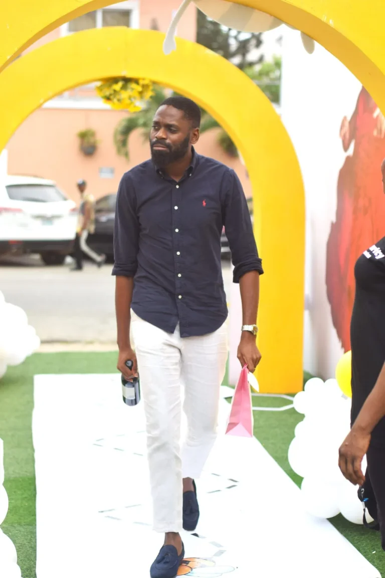 Bearded man in dark shirt and white trousers walks through a yellow arch carrying a gift bag.