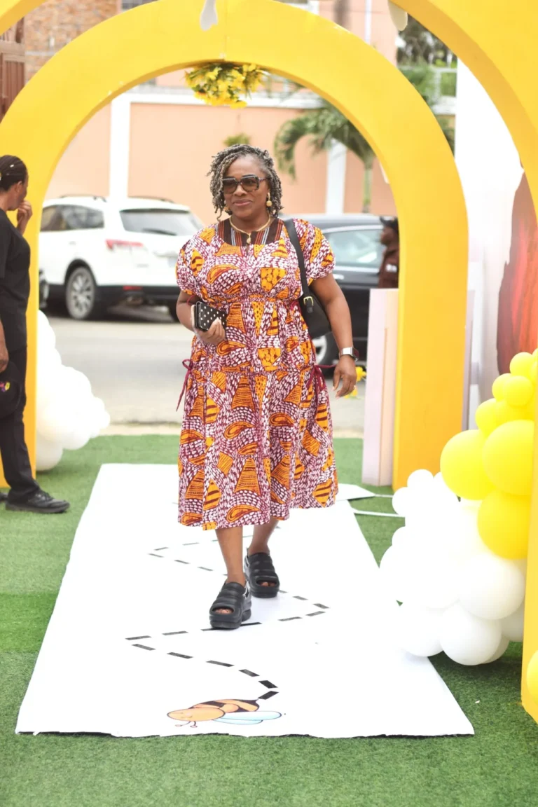 Woman in patterned African-print dress walks through a yellow archway with a clutch bag.