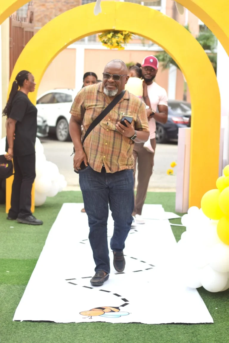 Older man in checked shirt and sunglasses walks holding a phone through a yellow arch.