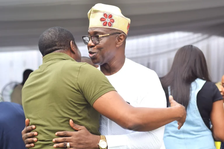 Two men in traditional and casual attire share a warm embrace at an indoor gathering.