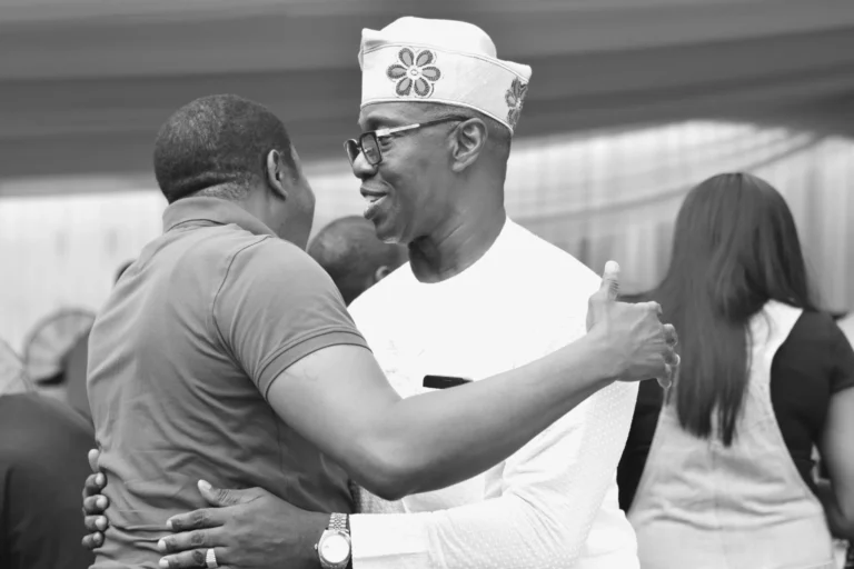 Two men embrace in greeting at an indoor event, rendered in black and white.