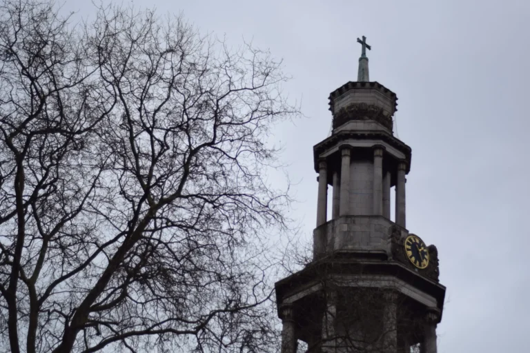 Stone church clock tower with weathercock cross, framed against grey winter sky and bare branches.
