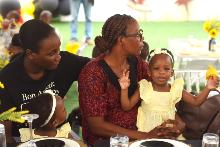 Two women seated at a decorated table holding young children in pale yellow dresses.