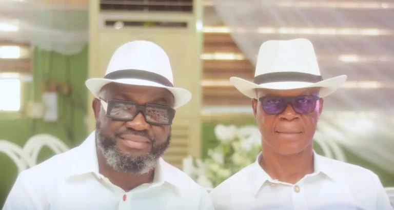 Two men in matching white fedoras and sunglasses pose side by side indoors.
