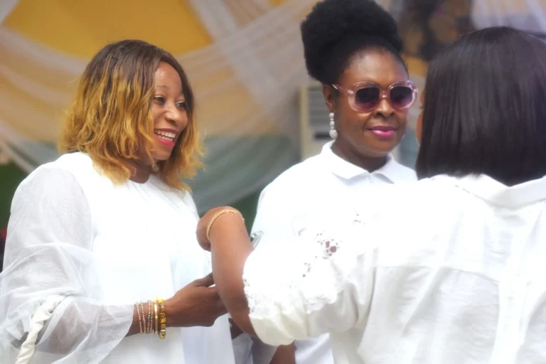 Three women in white outfits chat warmly beneath a yellow and white marquee.
