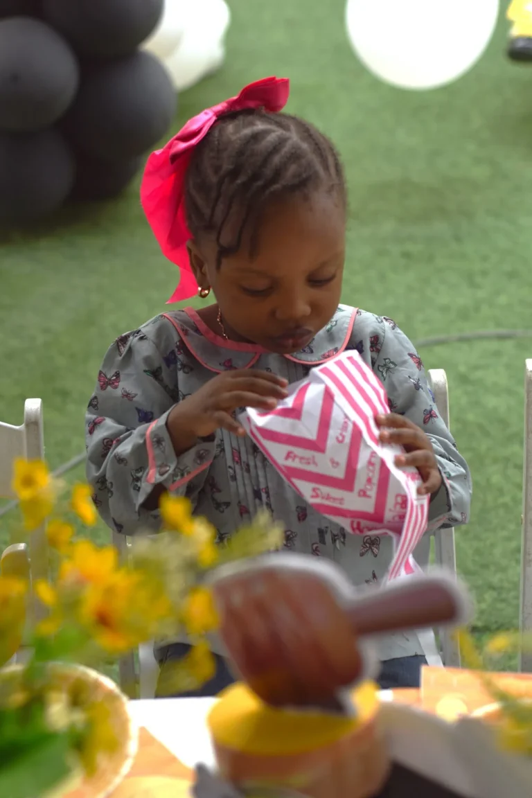 Young girl with red bow opens a striped popcorn packet at a party table.