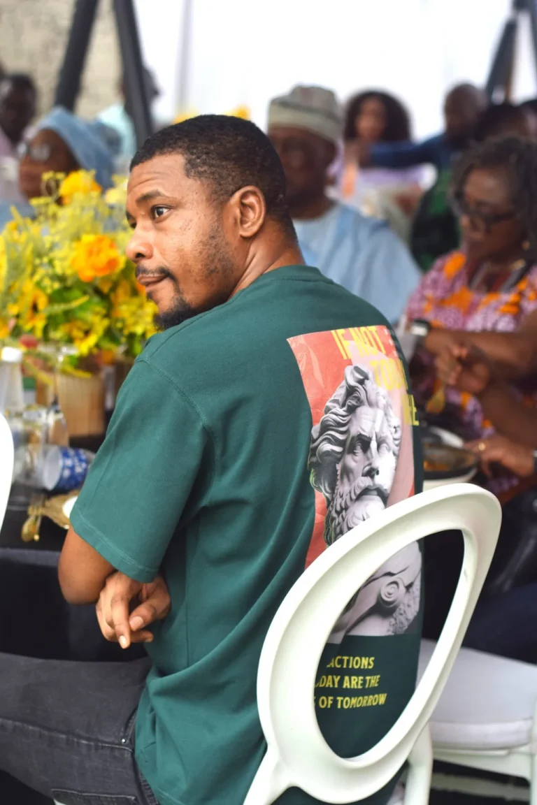 Man in green slogan T-shirt seated on a white chair amongst other guests outdoors.