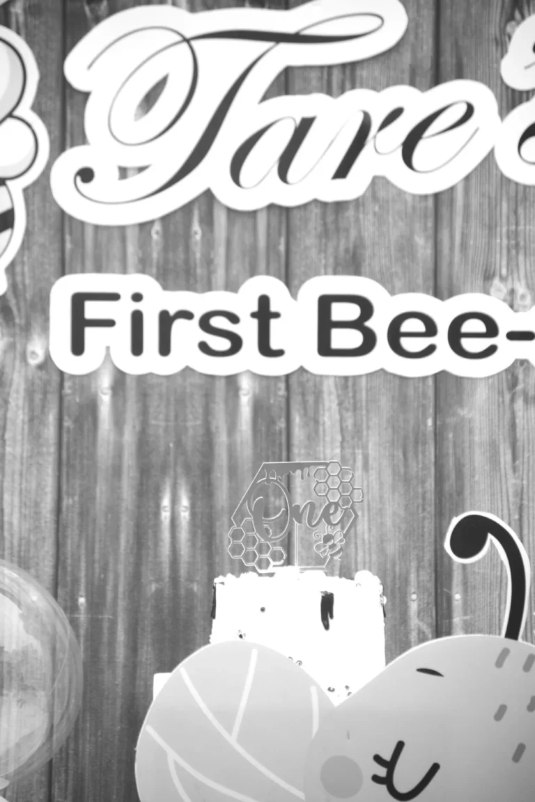 Black-and-white close-up of a wooden party backdrop with First Bee-day cake topper signage.