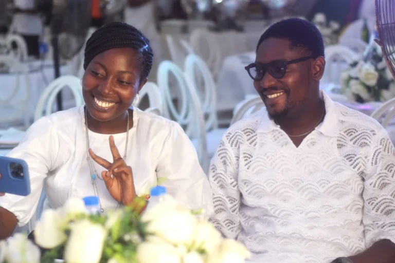 Smiling young woman makes a peace sign beside a grinning man in lace shirt.
