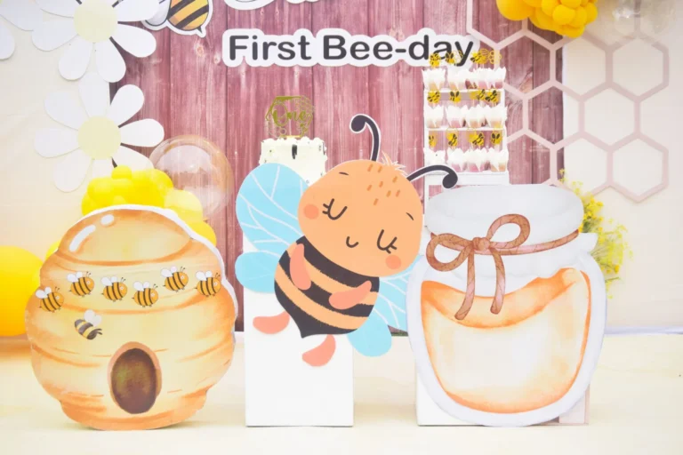 Cartoon bee, beehive and honey jar cut-outs arranged in front of a First Bee-day backdrop.