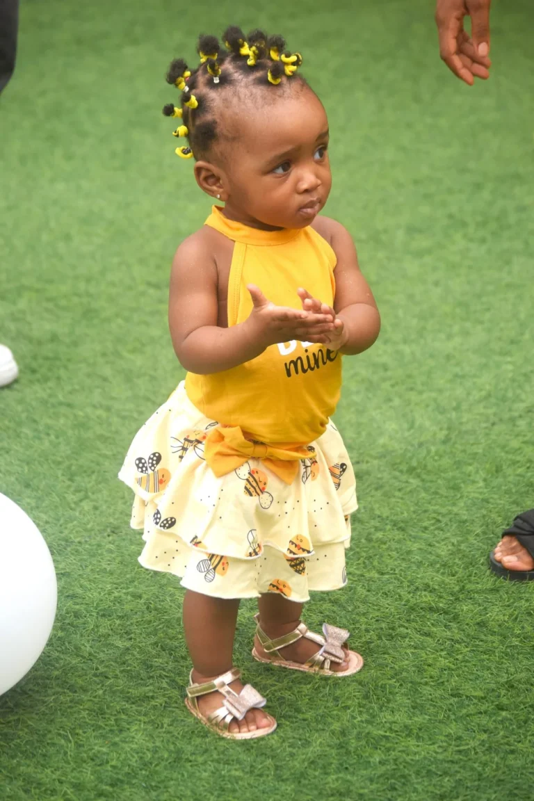 Toddler in yellow Bee Mine vest and printed skirt stands clapping on artificial grass.