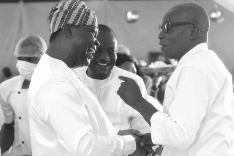 Two men shake hands and laugh together at a black-and-white indoor gathering.