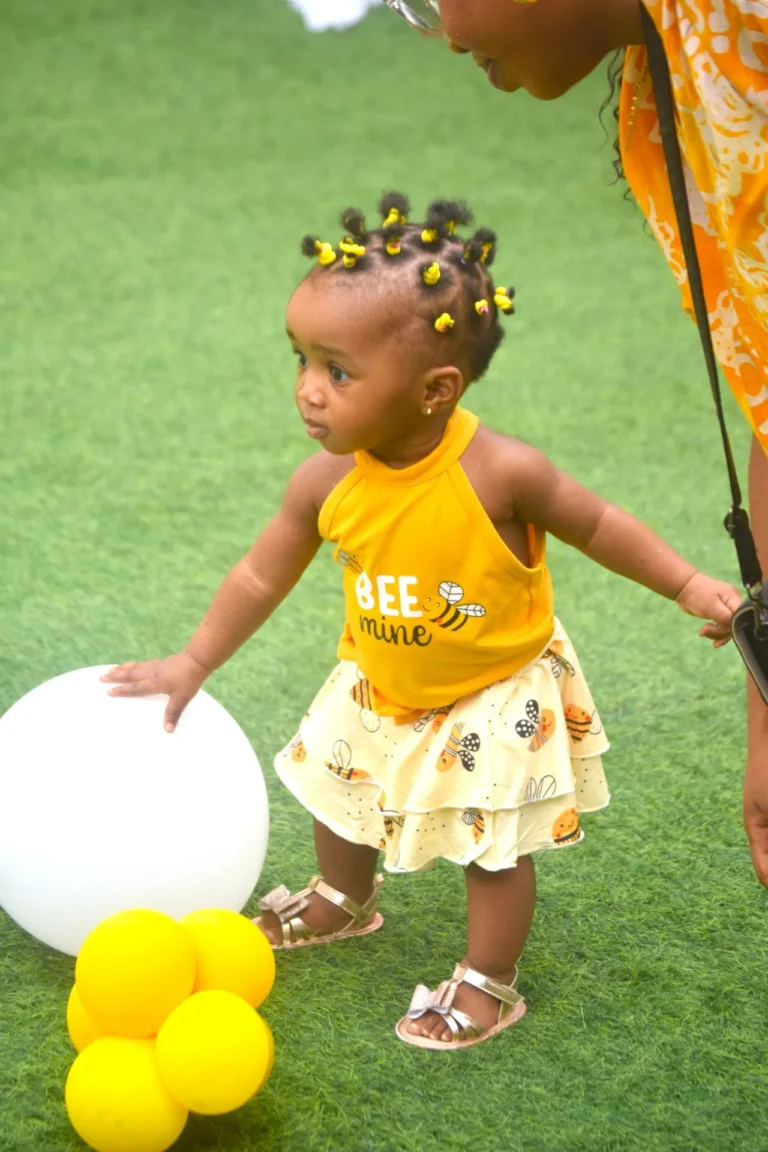 Toddler in yellow Bee Mine outfit reaches towards a large white ball on artificial grass.