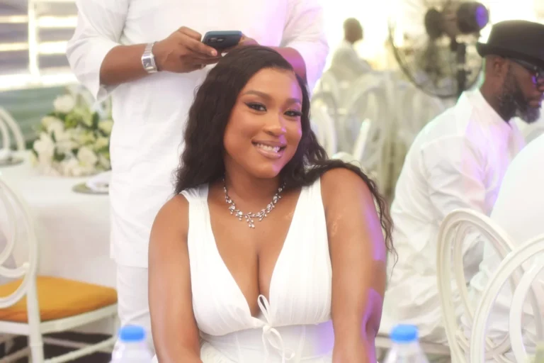 Young woman in white plunge dress smiles warmly at a busy reception table.