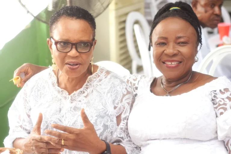 Two older women in white lace dresses smile together at an outdoor celebration.
